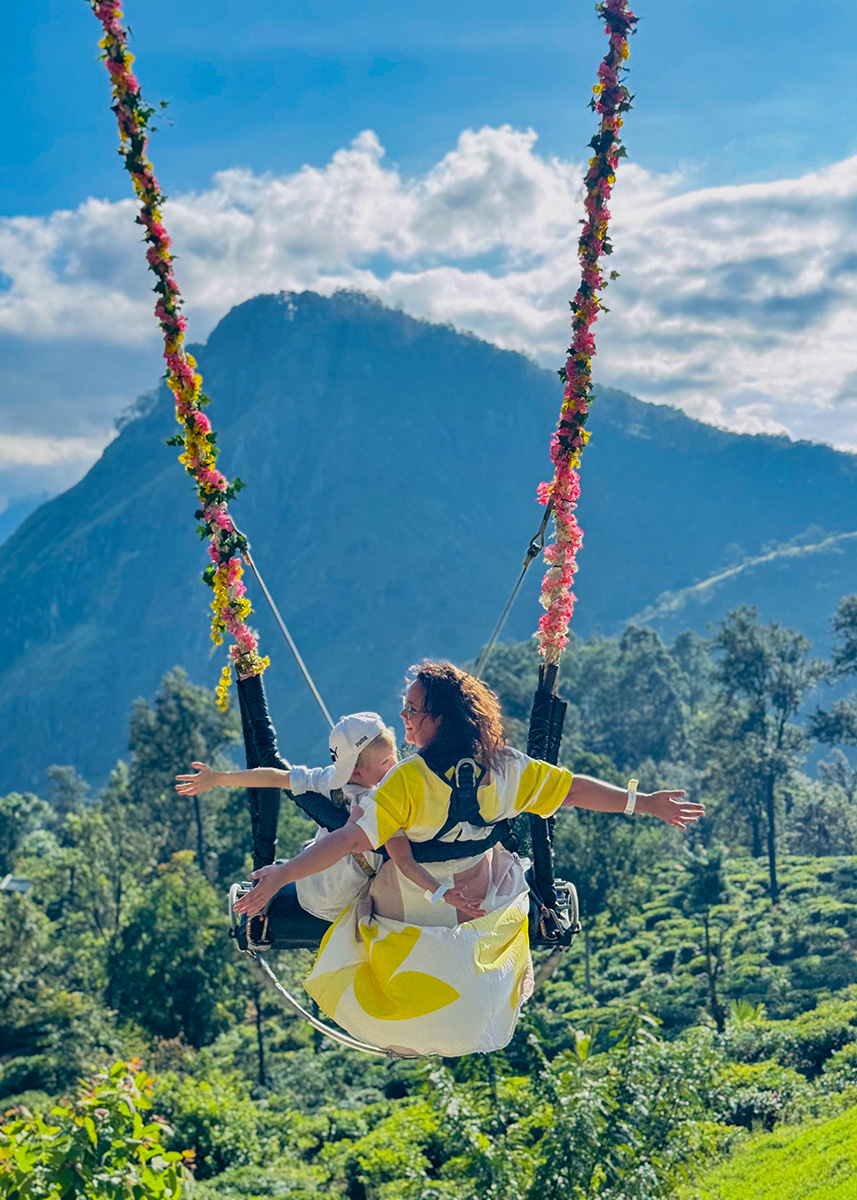 Mother and child on a flower-decorated swing in Ella, arms wide open with green hills and misty peaks behind them.