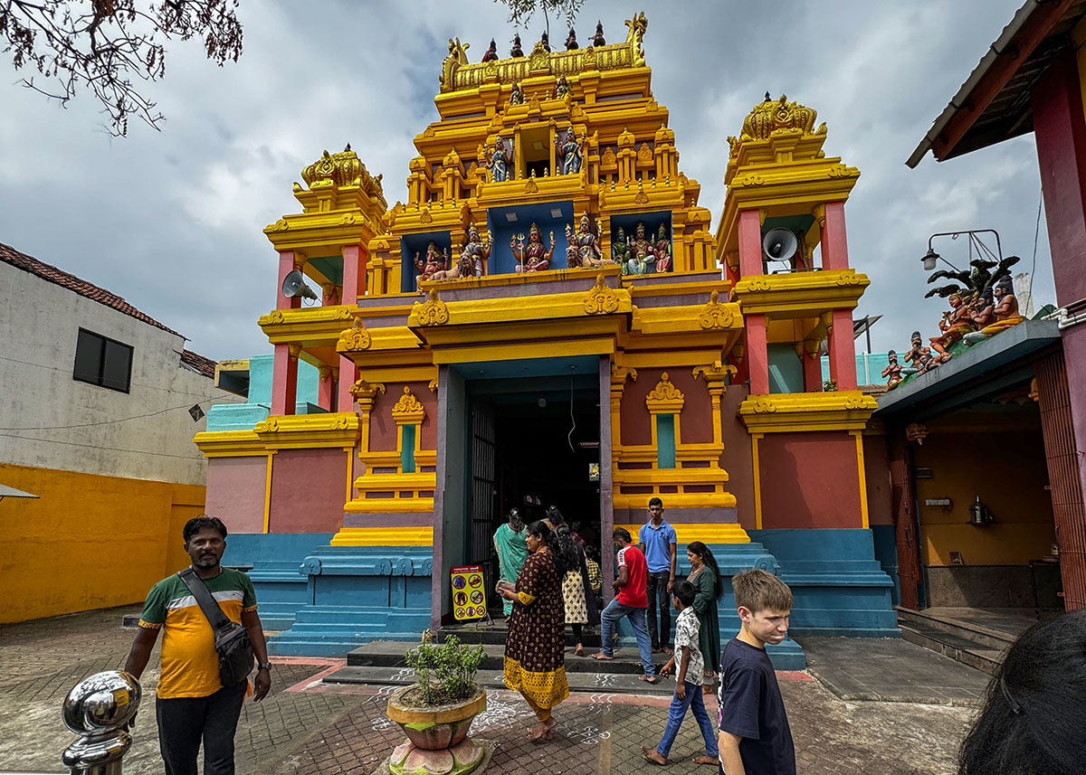 Visitors enter a vibrant yellow and red Hindu temple in Negombo, beneath moody skies, surrounded by locals.