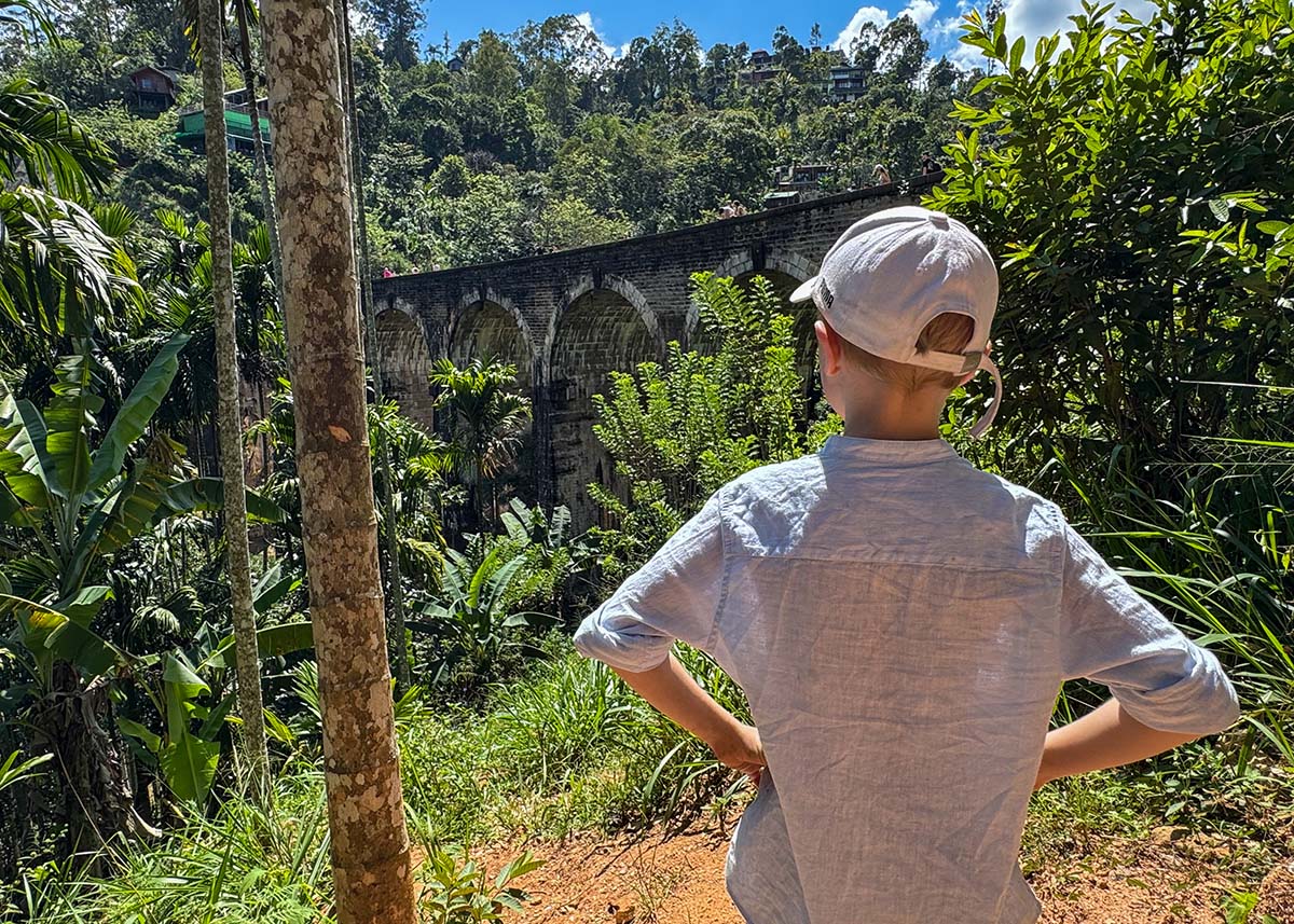 Child standing at a jungle viewpoint overlooking Nine Arches Bridge in Ella, Sri Lanka. The stone arches are partially visible through tropical trees. The weather is sunny and bright.
