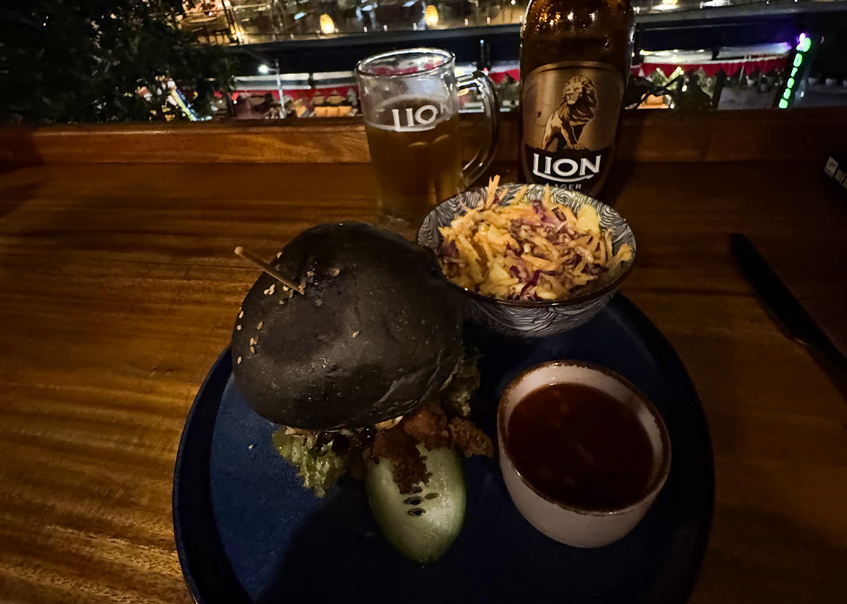 Burger served with fries, salad, and Lion beer at a restaurant in Ella, Sri Lanka. The table overlooks a lively street scene at night. The setting feels casual and family-friendly.