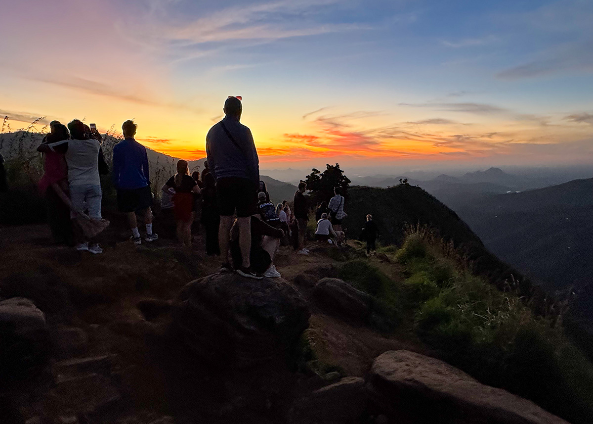 Crowds gathered at the summit of Little Adam’s Peak in Ella, Sri Lanka, watching sunrise. The sky glows orange and pink over rolling hills. Families and travelers stand on rocky viewpoints.