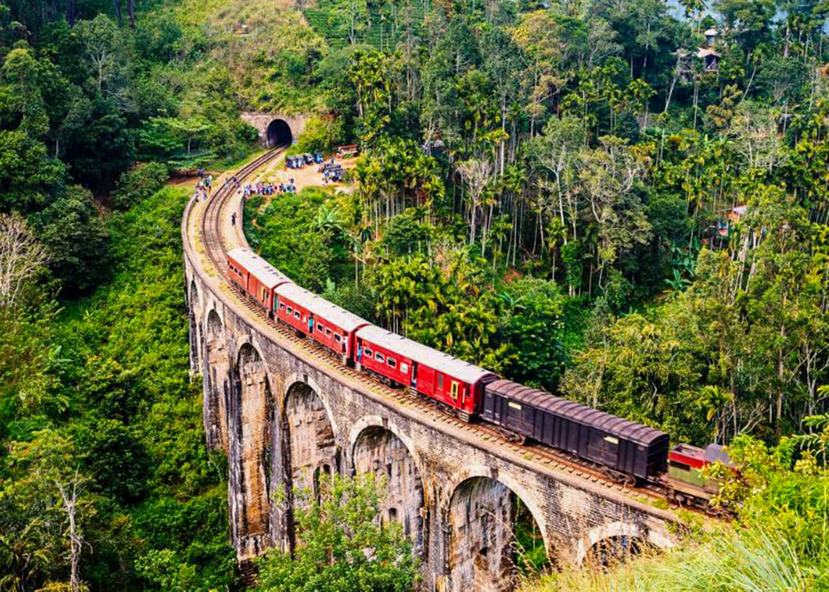 Red train crossing the iconic Nine Arches Bridge in Ella, Sri Lanka, surrounded by dense green jungle. Tourists gather near the tunnel entrance to watch the train pass. The weather is bright with clear visibility.