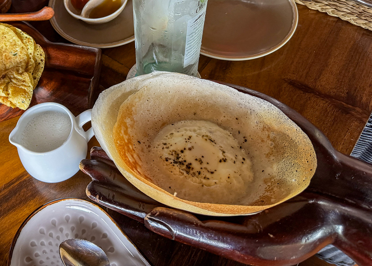 Traditional Sri Lankan hopper served with curry and sambal at a restaurant in Ella, Sri Lanka. The bowl-shaped pancake sits on a wooden tray with sauces and a drink nearby. The meal is presented in a casual dining setting.