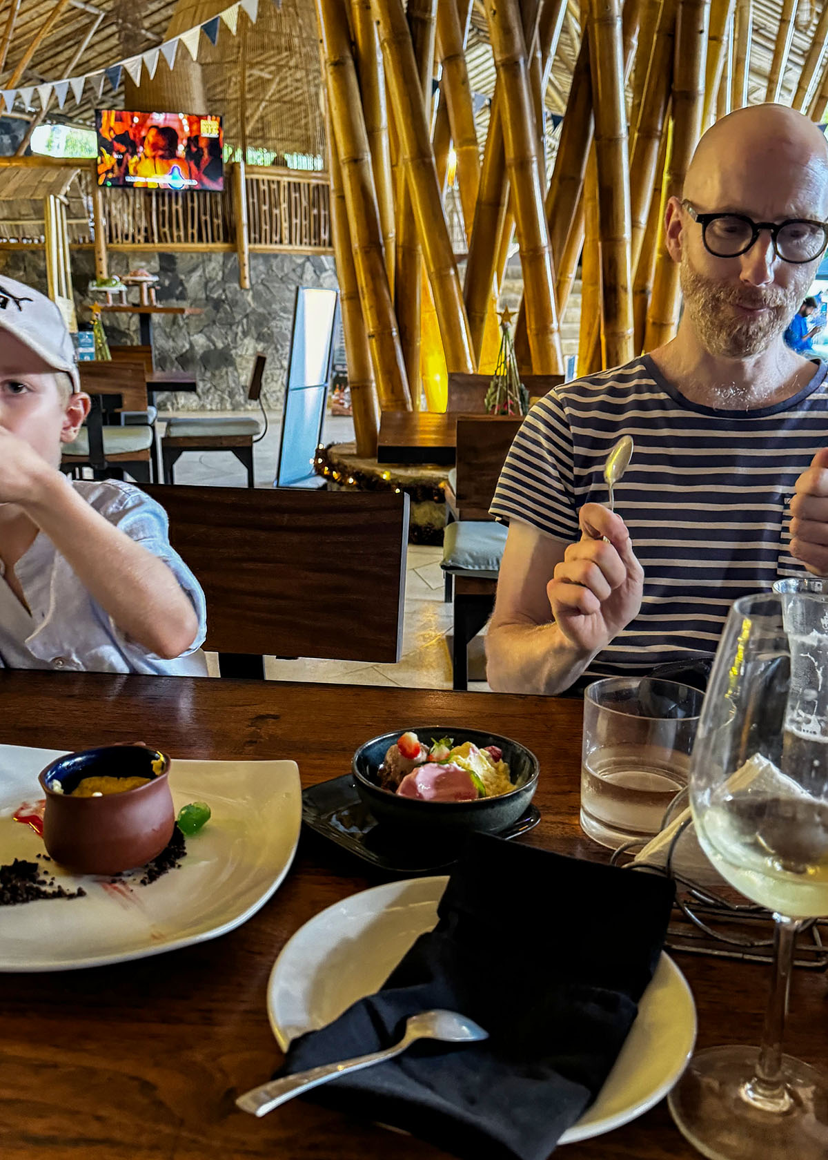 Man and child enjoying dessert at a bamboo-style restaurant in Ella, Sri Lanka. The table holds plated sweets and drinks under warm indoor lighting. The open-air setting features wooden furniture and a relaxed family atmosphere.