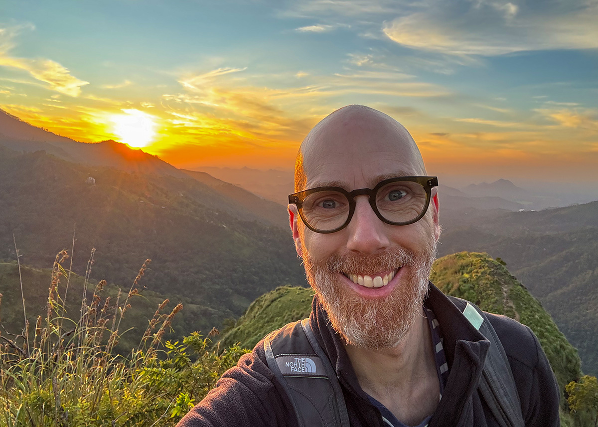 Man taking a selfie at sunrise on Little Adam’s Peak in Ella, Sri Lanka. The sun rises behind rolling hills, casting warm light across the landscape. The sky is clear with soft clouds.