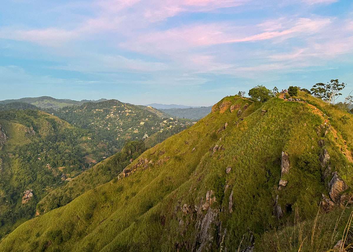 View of the grassy ridge leading to Little Adam’s Peak in Ella, Sri Lanka. Visitors walk along the narrow trail under soft morning light. The surrounding hills stretch into the distance.