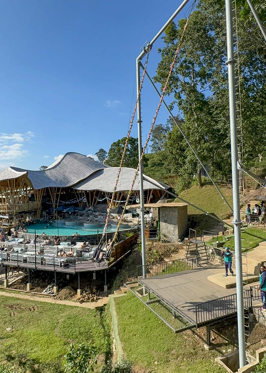 Large swing structure overlooking Ravana Pool Club near Ella, Sri Lanka. Visitors relax by the pool while others prepare to swing over the area. The weather is sunny with clear skies.