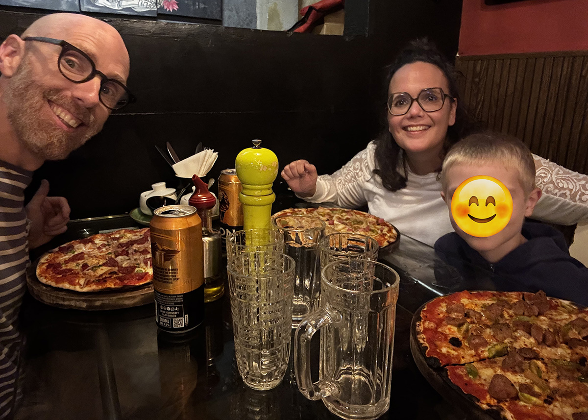 Family sitting at a restaurant in Ella, Sri Lanka, with large pizzas and drinks on the table. The indoor setting is dimly lit with a cozy evening atmosphere. Everyone appears relaxed and enjoying a casual meal.