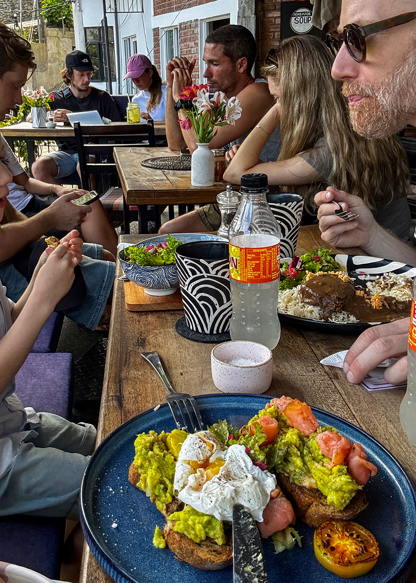 Family enjoying lunch at a popular café in Ella, Sri Lanka. Avocado toast, poached eggs, and drinks are served on a wooden table. Other travelers dine nearby in a relaxed daytime setting.