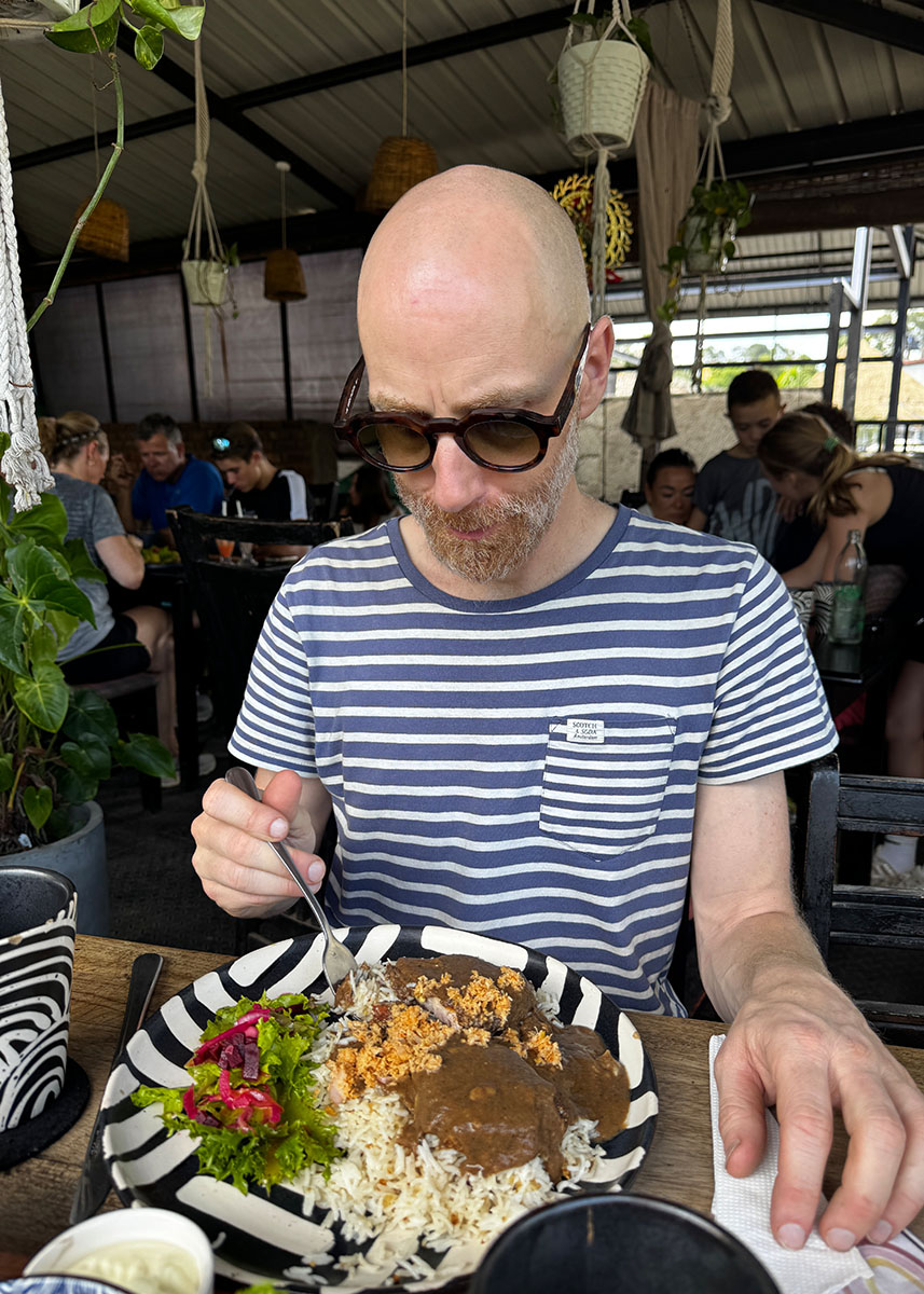 Man eating a large plate of rice and curry at a busy café in Ella, Sri Lanka. The dish includes curry sauce, coconut sambal, and salad on a patterned plate. Other travelers sit in the background.
