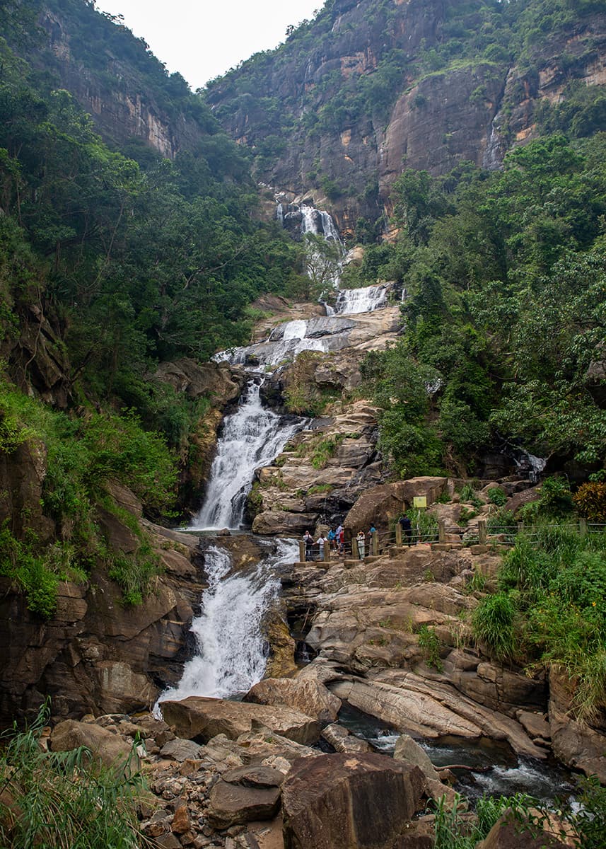 Wide view of Ravana Falls in Ella, Sri Lanka, showing multiple tiers of cascading water and visitors on viewing platforms. The surrounding jungle is lush. The weather is overcast.