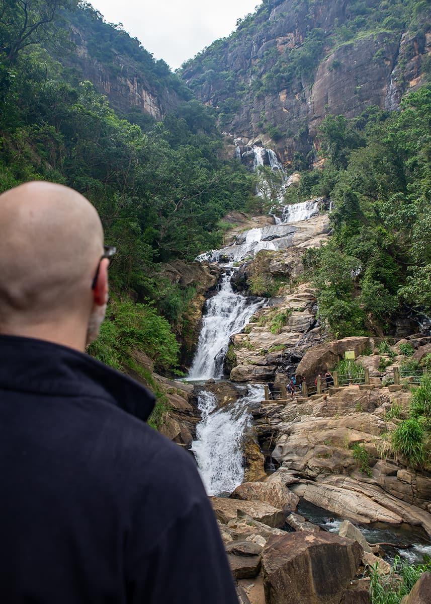 Man standing in front of Ravana Falls in Ella, Sri Lanka, watching the cascading water over rocky cliffs. The waterfall flows through dense greenery. The sky is cloudy.