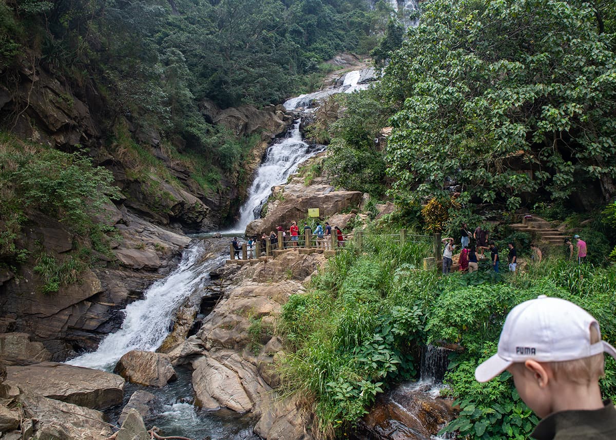 Child looking toward Ravana Falls in Ella, Sri Lanka, from a nearby path. Visitors stand on a platform beside the waterfall. The scene is shaded with cloudy skies.