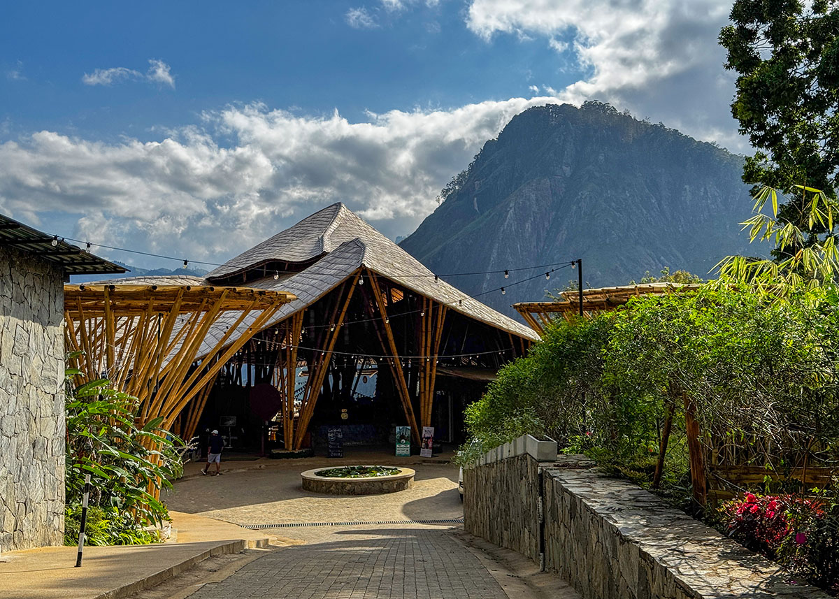 Modern bamboo-style entrance to Ravana Pool Club near Ella, Sri Lanka. The building sits against a mountainous backdrop under partly cloudy skies. A paved walkway leads into the complex.