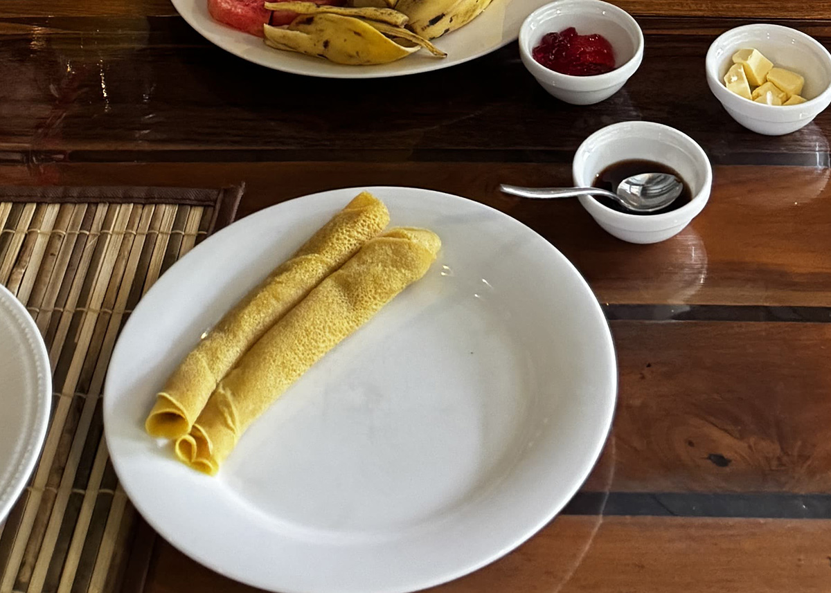 Plate of rolled pancakes served with fruit, butter, and jam at a breakfast spot in Ella, Sri Lanka. Small bowls of condiments sit on a wooden table. The setting is bright and relaxed.