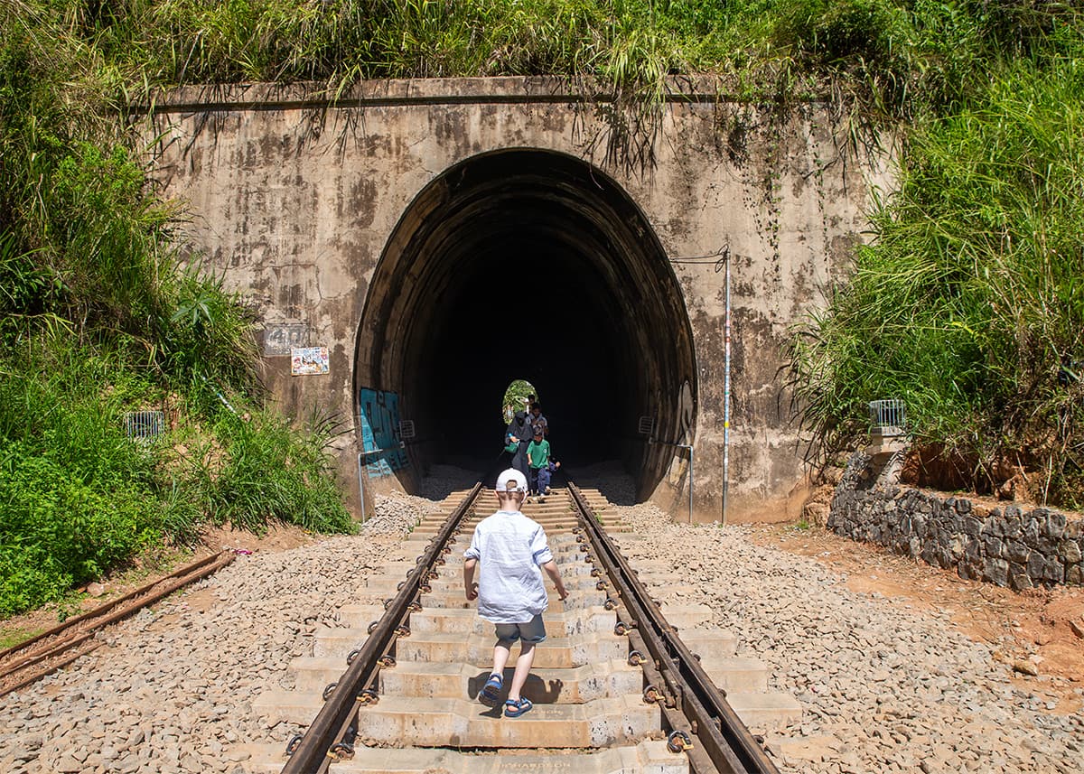 Child walking along railway tracks toward a dark tunnel near Ella, Sri Lanka. Other visitors stand inside the tunnel entrance, surrounded by greenery and rocky hillsides. The weather is sunny and warm.