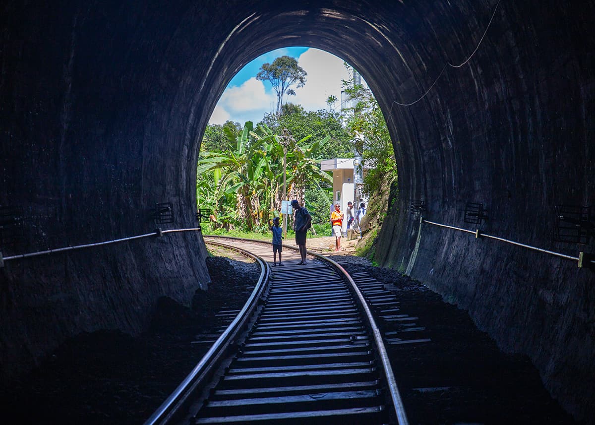 View from inside a railway tunnel near Ella, Sri Lanka, looking out toward bright greenery. A few visitors stand near the tunnel exit on the tracks. The scene contrasts dark interior with sunny outdoors.