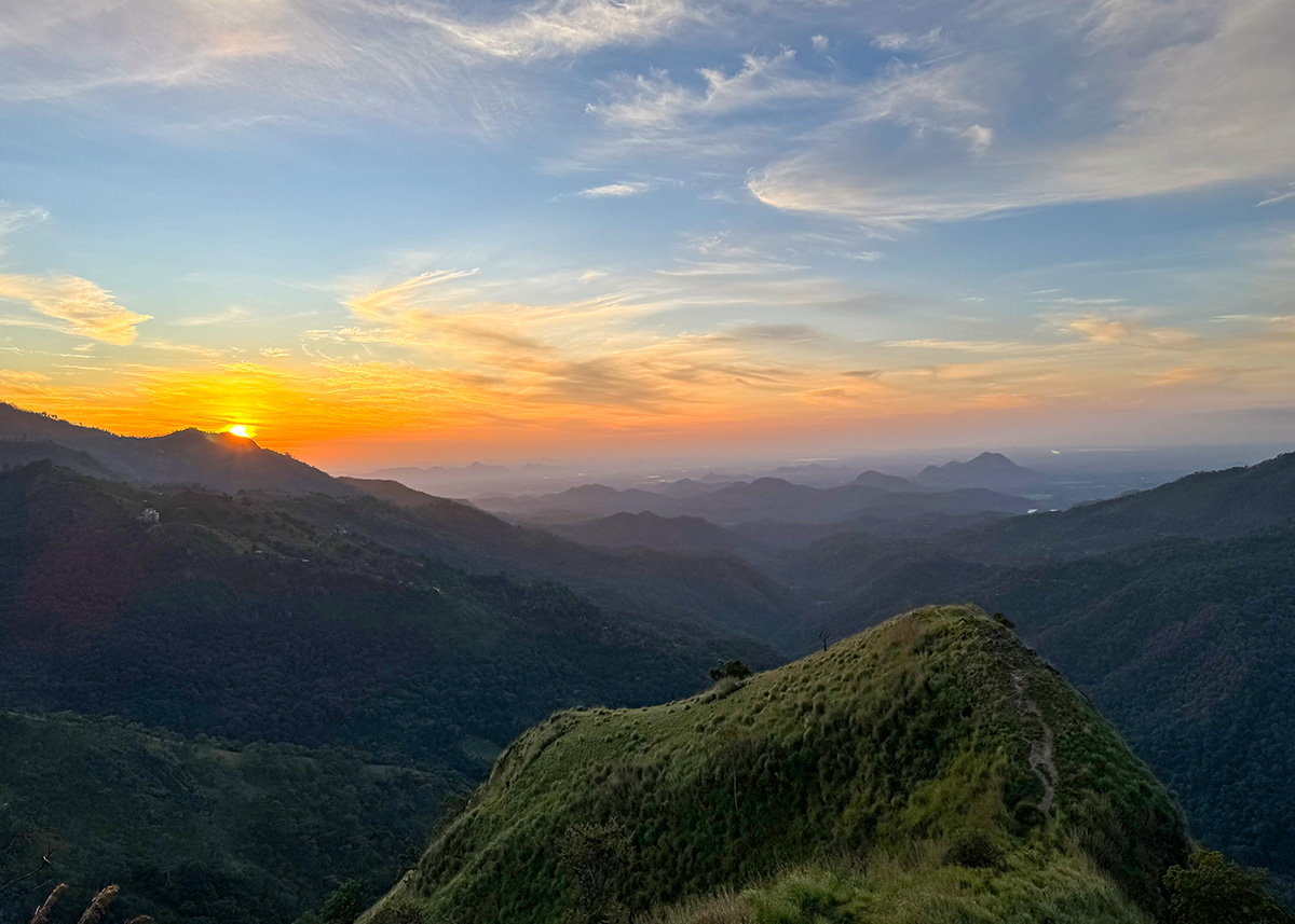 Golden sunrise over the mountains of Ella, Sri Lanka, viewed from Little Adam’s Peak. Layers of hills fade into the horizon under a colorful sky. The scene is calm and scenic.