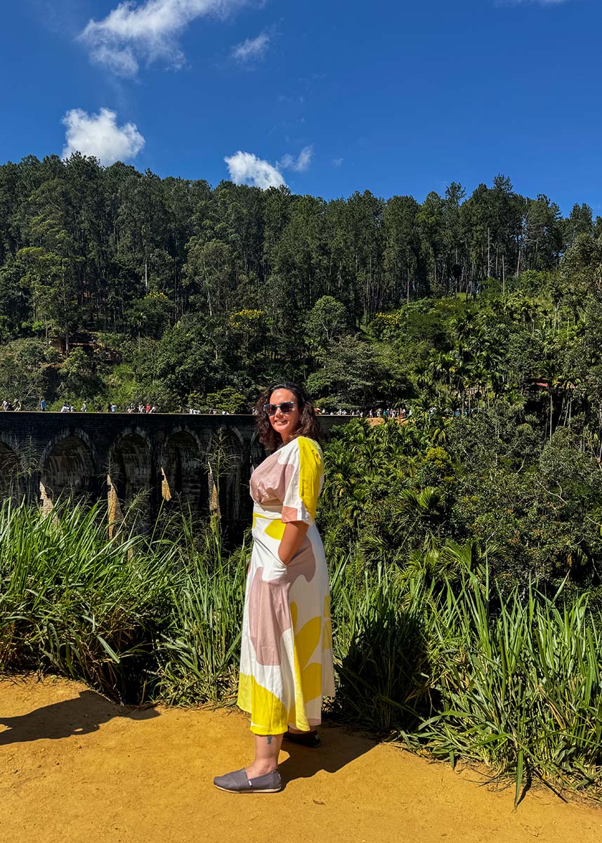 Woman posing at a viewpoint above Nine Arches Bridge in Ella, Sri Lanka. The sunny weather highlights the green jungle and stone arches below. Tourists can be seen walking across the bridge.