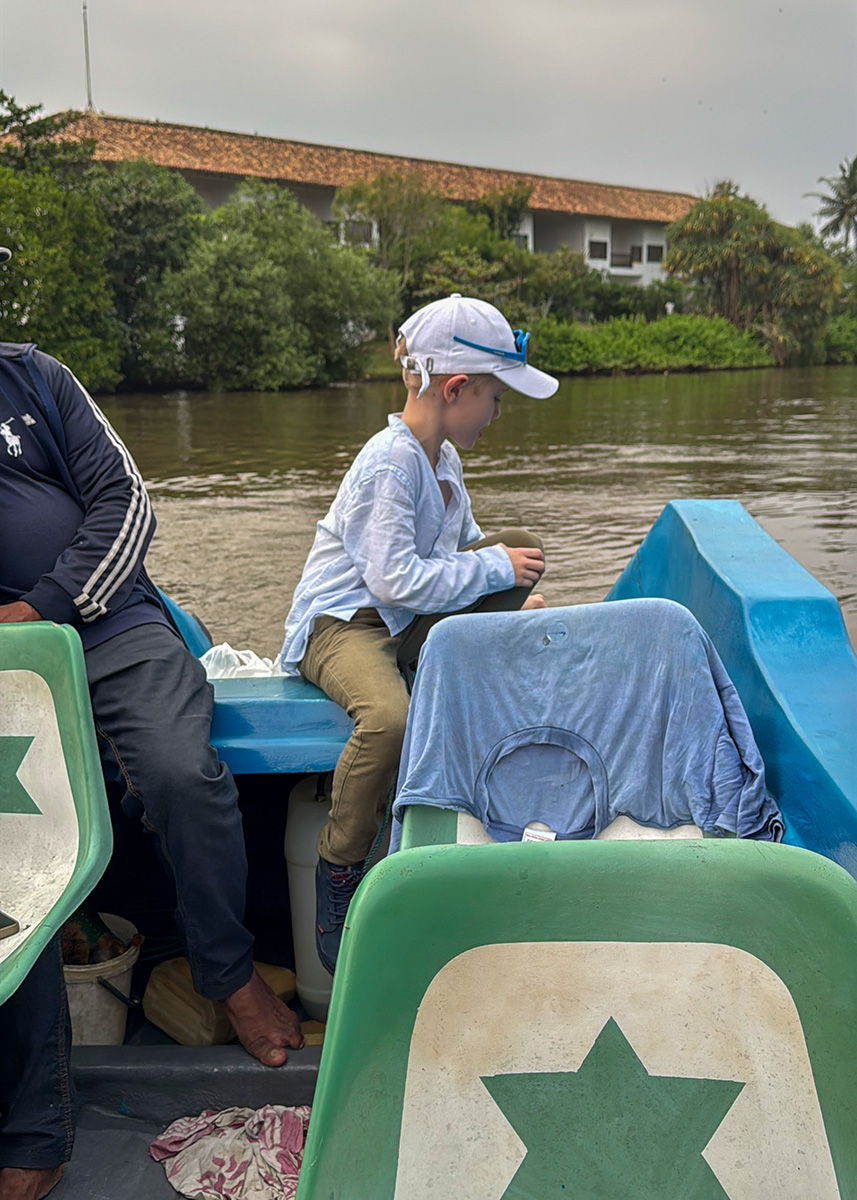 Child sitting in a small boat during a lagoon tour in Negombo with houses and greenery along the water.