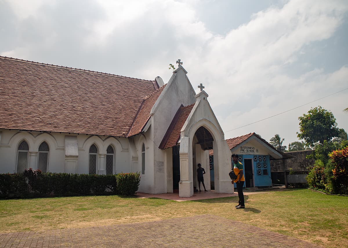White church with red roof in Negombo, Sri Lanka, surrounded by grass and palm trees. A man stands near the entrance. The weather is sunny and warm.
