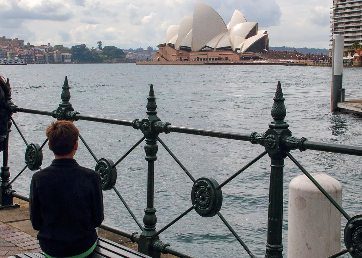 Young boy sitting beside Sydney Harbour looking toward the Sydney Opera House across the water. Decorative black iron railings frame the harbor view on a cloudy afternoon.