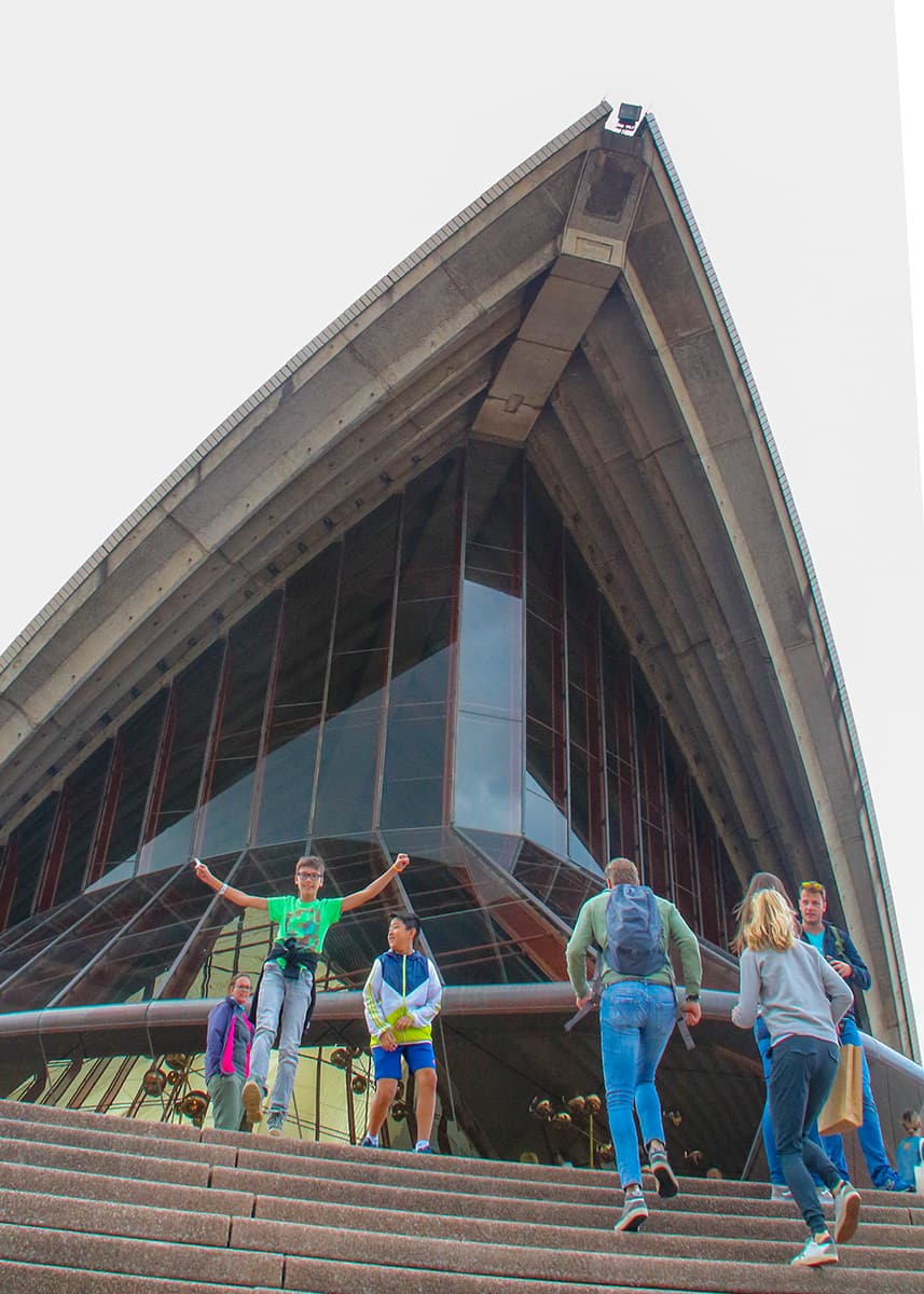 People walk up the wide steps outside the Sydney Opera House on a cloudy day in Sydney Harbour. The sharp concrete sails and glass facade tower overhead while a boy in a bright green shirt jumps excitedly near the entrance,