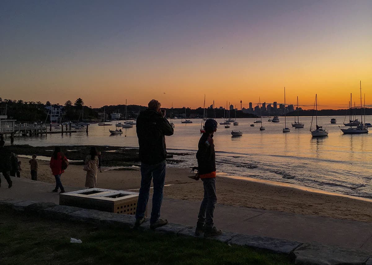 Family standing along the shoreline at sunset in Sydney Harbour while sailboats float across calm water. The Sydney skyline appears in silhouette against an orange and purple evening sky.