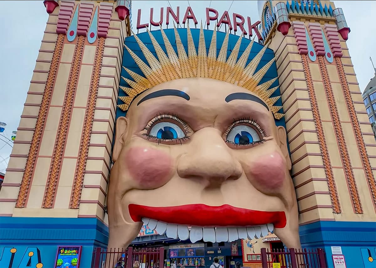 Entrance to Luna Park Sydney featuring the famous giant smiling face and colorful Art Deco towers. The amusement park entrance stands beneath an overcast sky with visitors walking below the sign reading “LUNA PARK.”