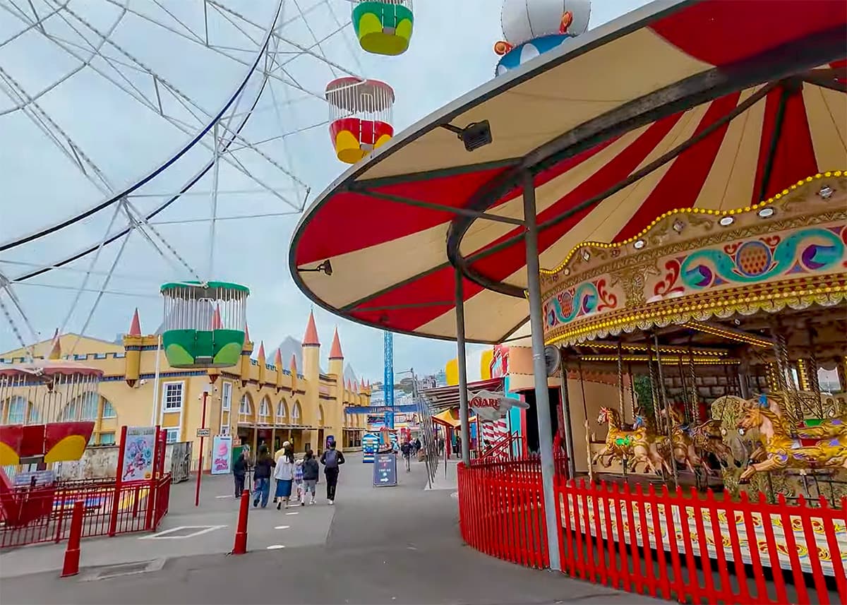 Colorful rides and carousel at Luna Park Sydney on a cloudy day. Red fencing, hanging swing ride seats and families walking through the amusement park entrance area fill the scene.