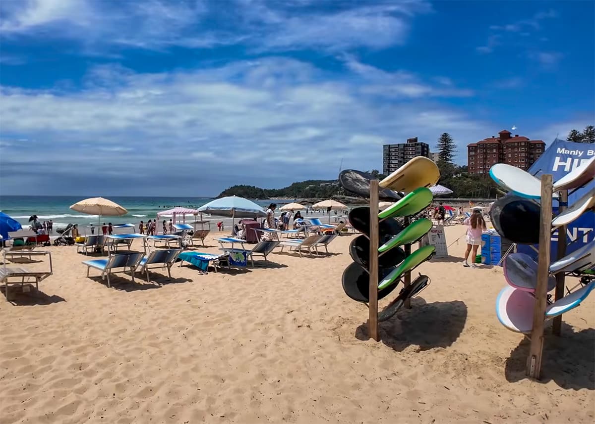 Busy beach scene at Manly Beach Sydney with surfboards stored upright in the sand beside beach umbrellas and lounge chairs. Families relax near the shoreline under a partly cloudy blue sky.