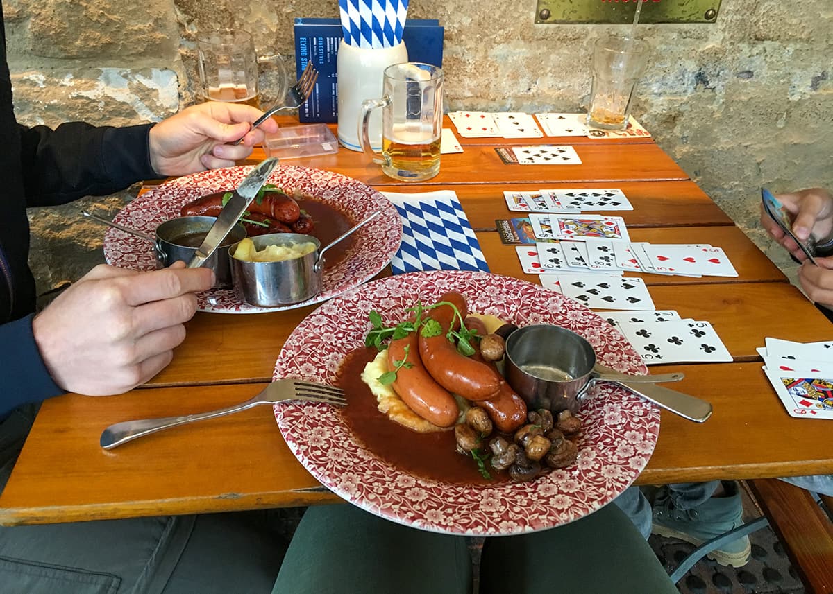Plates of sausages, mashed potatoes and mushrooms served at a restaurant in Sydney while a family plays cards at the wooden table filled with drinks, food and scattered playing cards.