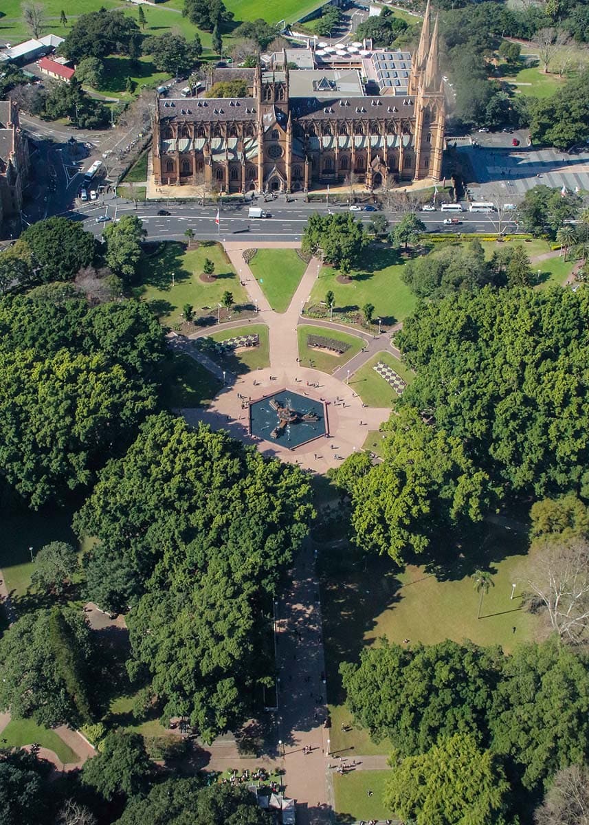 Aerial view of St Mary’s Cathedral and Hyde Park in Sydney on a clear sunny day. Tree lined walking paths and the central fountain stretch through the large city park below.