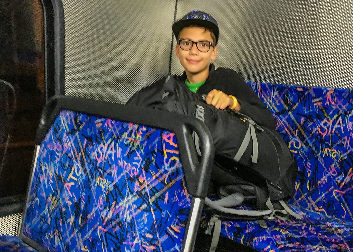 Young boy wearing glasses and a baseball cap sits on a Sydney public bus holding a backpack on his lap. Bright patterned bus seats and dim evening lighting give the scene a relaxed family travel feel.