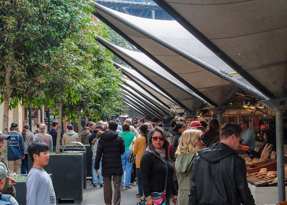 Crowded outdoor market lane at The Rocks Market in Sydney filled with shoppers browsing vendor stalls. Covered awnings and leafy trees line the busy pedestrian walkway on a cool cloudy day.