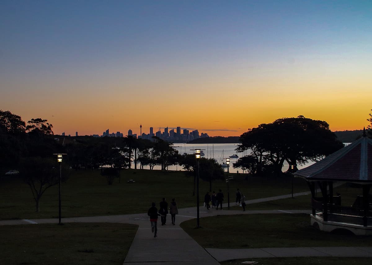 Sunset view across Robertson Park toward Sydney Harbour and the city skyline at dusk. Walking paths, scattered trees and small groups of people lead toward the glowing orange horizon.