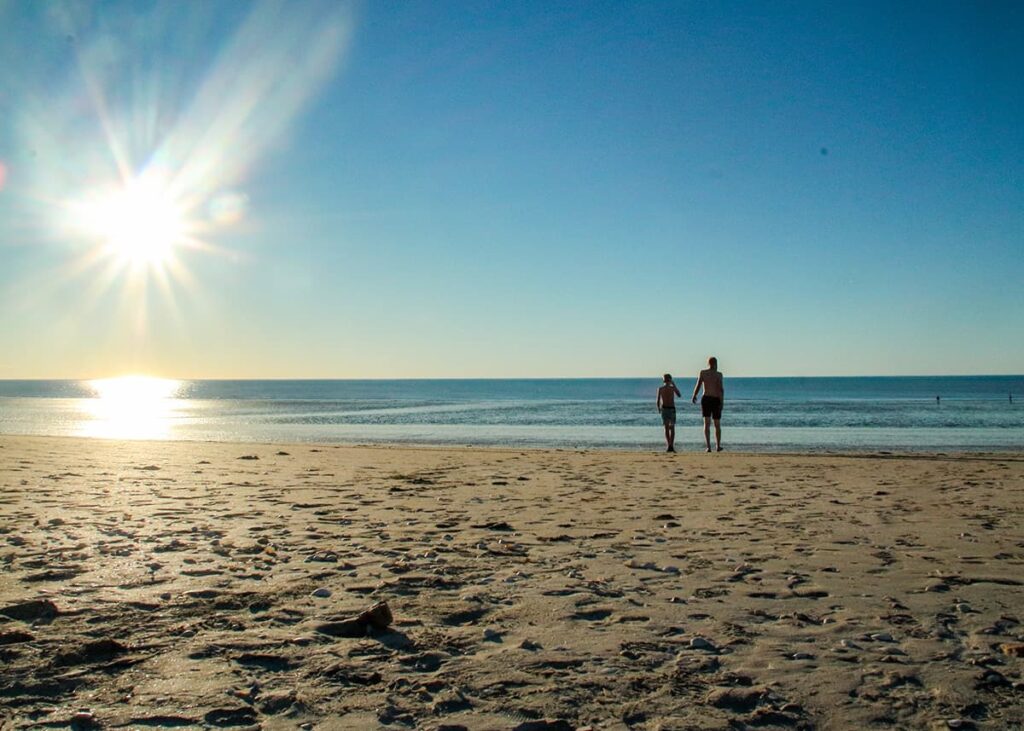 A father and son are walking on the beach towards the sea, while the sun is slowly setting in the background