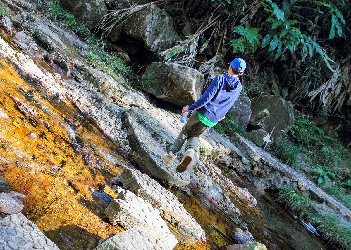 A child carefully crosses rocky terrain beside a shallow stream during a hike in the Cardamom Mountains, Cambodia. Surrounded by tropical vegetation, large boulders, and clear flowing water, the scene captures a family adventure in the rainforest. The sunny weather makes the colorful rocks and stream stand out against the lush jungle backdrop.