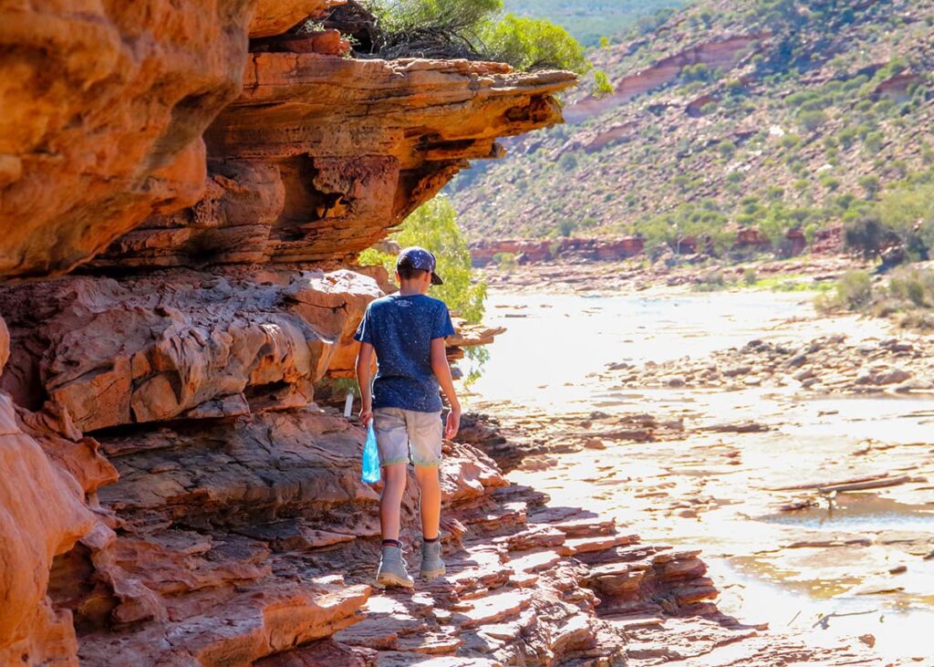 Child walking beside layered red sandstone cliffs at Kalbarri National Park in Western Australia on a bright sunny day. The rocky riverbank trail follows the Murchison River through the dramatic gorge landscape.