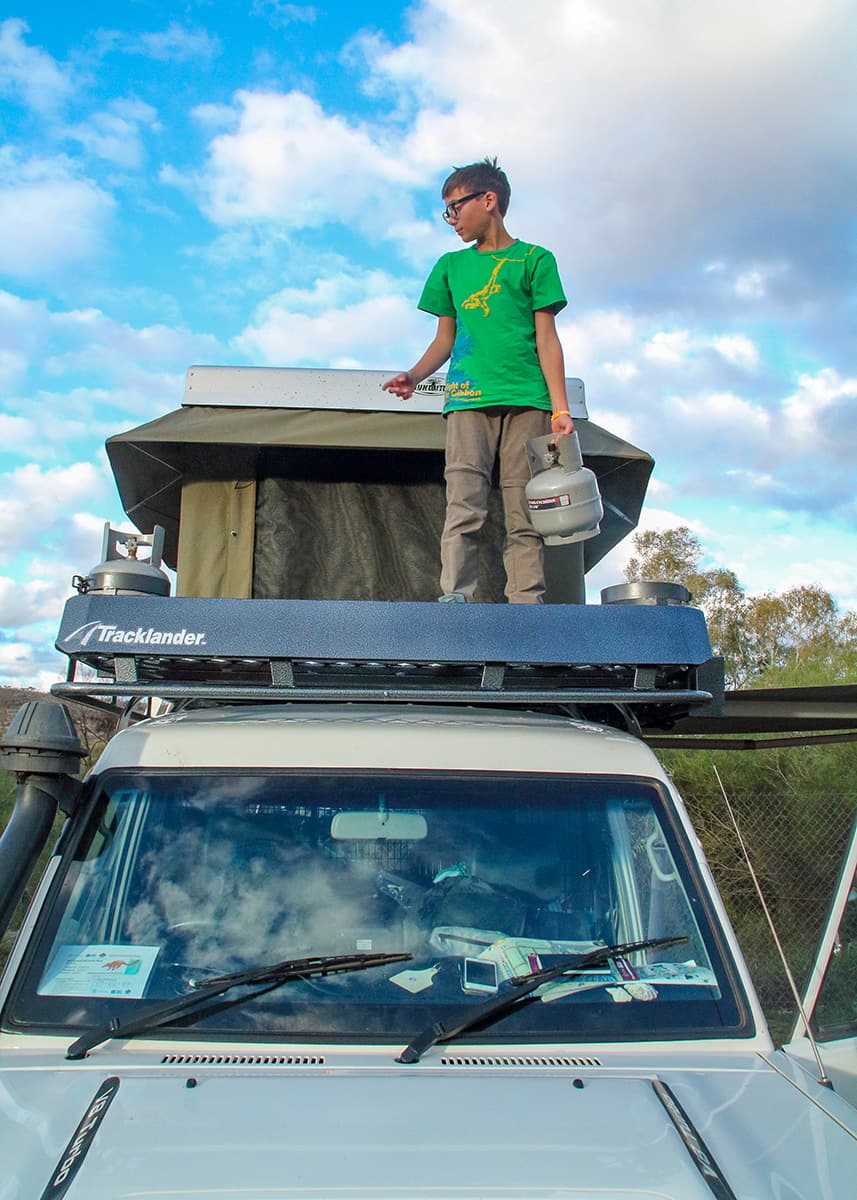 Child standing on the rooftop tent platform of a four wheel drive camper at a campsite in Tom Price. Puffy clouds fill the sky as he holds a gas bottle.