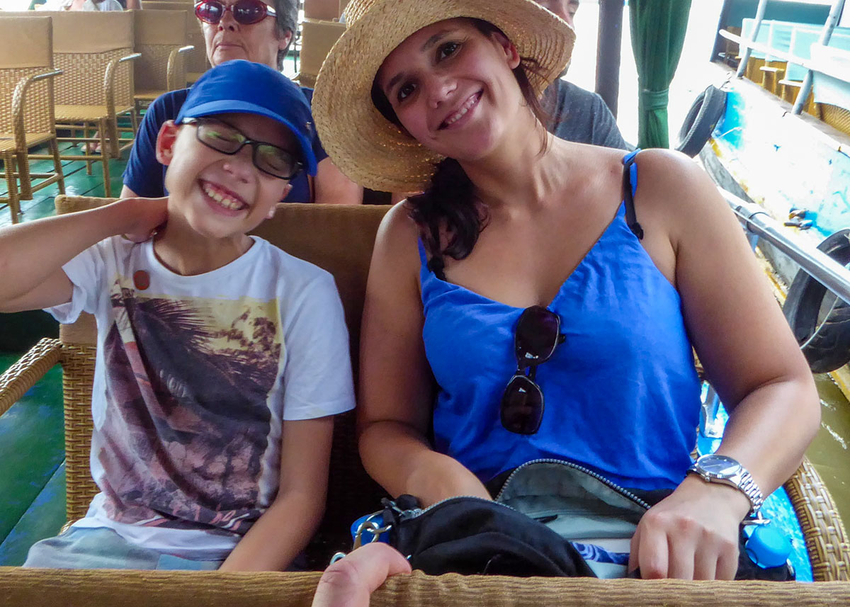 A smiling mother and child sit aboard a riverboat during an Irrawaddy dolphin tour near Kratie, Cambodia. The sunny weather and open-air seating create a relaxed atmosphere as families travel along the Mekong River searching for dolphins. Boat tours are one of the most popular family activities in the area.