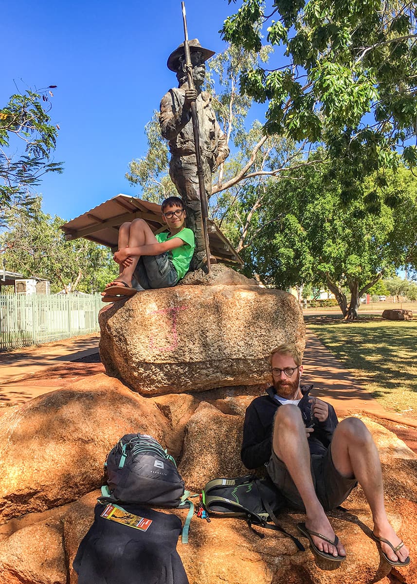 Child and parent resting beside the statue of Jack Jugarie in Halls Creek, Western Australia. Large granite boulders and shady trees surround the monument on a sunny day.