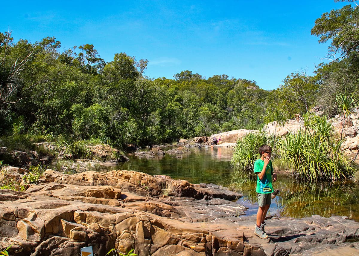 Child standing on rocky ledges beside the natural pools at Maguk in Kakadu National Park, Northern Territory. Clear water winds through the tropical landscape beneath a bright blue sky.