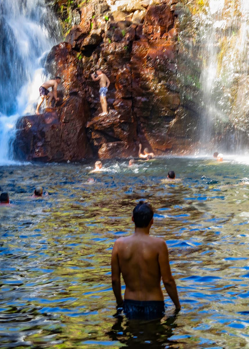 Visitors swimming beneath the cascading waters of Florence Falls in Litchfield National Park, Northern Territory. Children and adults enjoy the cool plunge pool surrounded by wet rock walls and tropical forest.