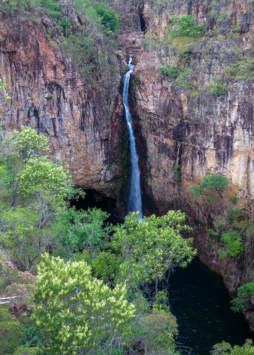 Tolmer Falls plunging into a dark pool within Litchfield National Park in the Northern Territory. Steep rocky cliffs and green vegetation frame the tall waterfall on a warm day.