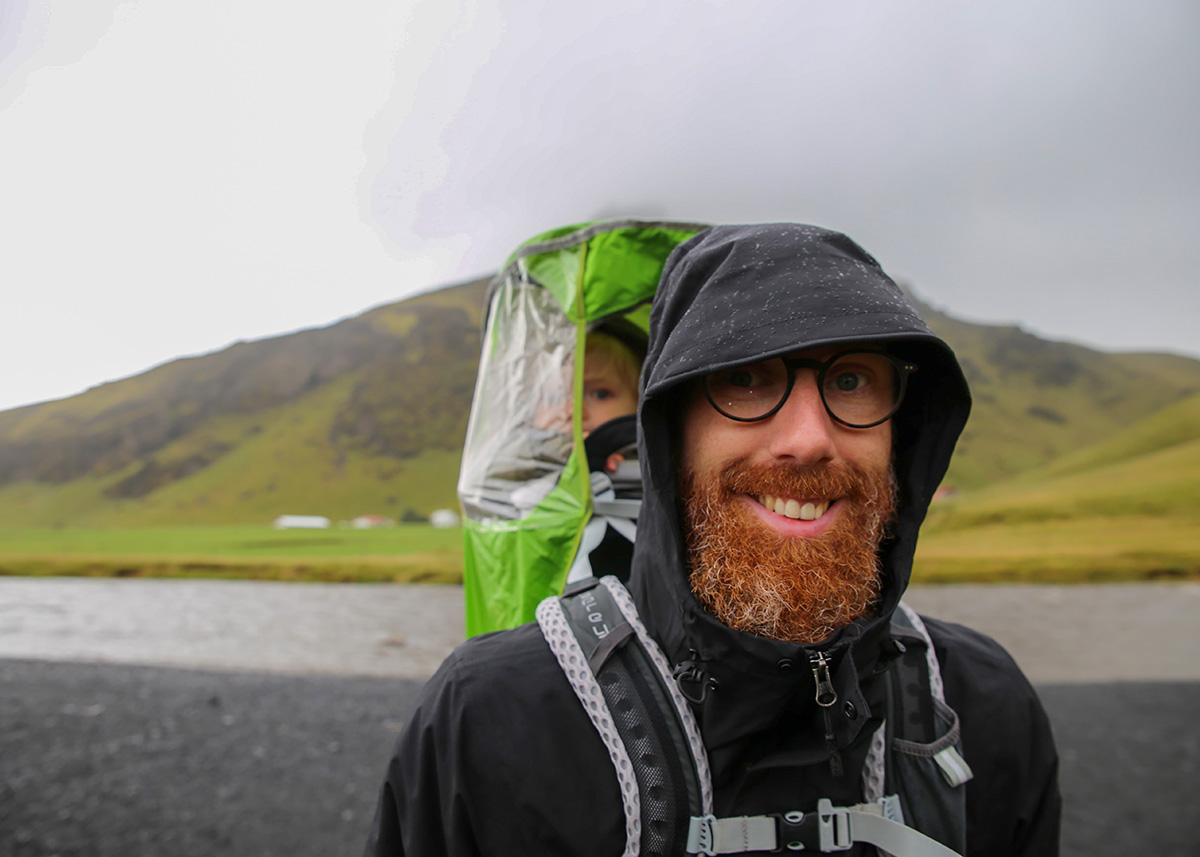 A father carries his toddler in a back carrier covered with a rain cover. They're standing on the black sand beach leading up to Skogafoss waterfall in Iceland